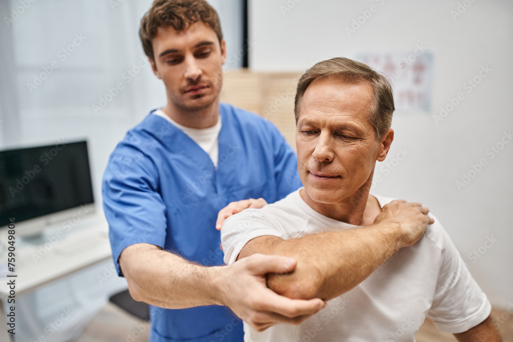good looking patient in casual attire stretching his muscles with help of his handsome doctor good looking patient in casual attire stretching his muscles with help of his handsome doctor