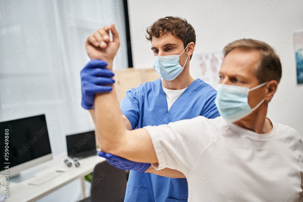 Obraz premium hardworking doctor in medical gloves and mask rehabilitating his mature patient in hospital
