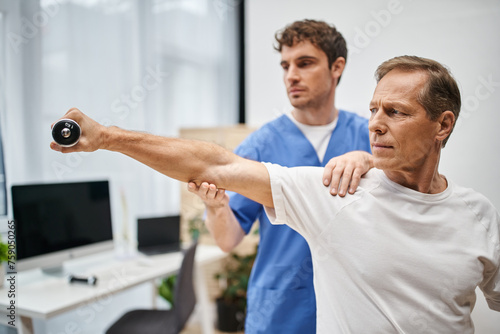 hardworking doctor in blue robe helping his mature patient with dumbbell during rehabilitation