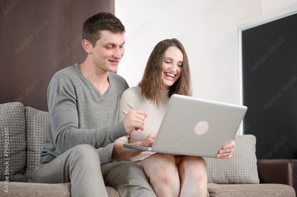 Happy young couple talking, browsing online website, shopping, using laptop while sitting on couch at home. Concept of technology, communication, spending weekends with family, family budget planning Happy young couple talking, browsing online website, shopping, using laptop while sitting on couch at home. Concept of technology, communication, spending weekends with family, family budget planning