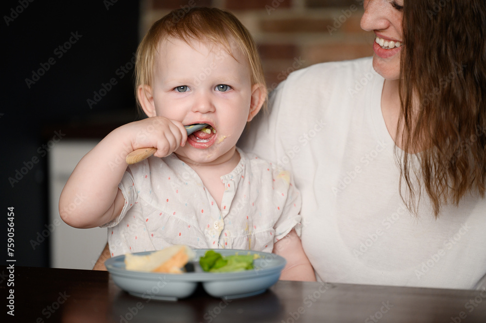 Child learns to eat independently, sitting in his mother arms in kitchen. Toddler independently eats mixture of mashed potatoes from plate with spoon. Concept of self-feeding, healthy eating, weaning. Child learns to eat independently, sitting in his mother arms in kitchen. Toddler independently eats mixture of mashed potatoes from plate with spoon. Concept of self-feeding, healthy eating, weaning.