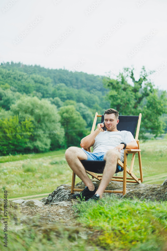 Cute smiling man on folding wooden chair sitting in nature and resting. The concept of recreation in nature alone.