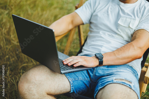 A man on a folding wooden chair sits in nature and works remotely online on a laptop. The concept of recreation in nature alone.