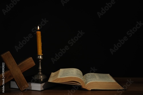 Church candle, Bible and cross on wooden table against black background, space for text
