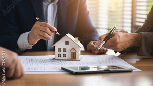 Small model house on a desk with two people in the background, suggesting a discussion, likely about property or finance, with pens and documents indicating a business environment.