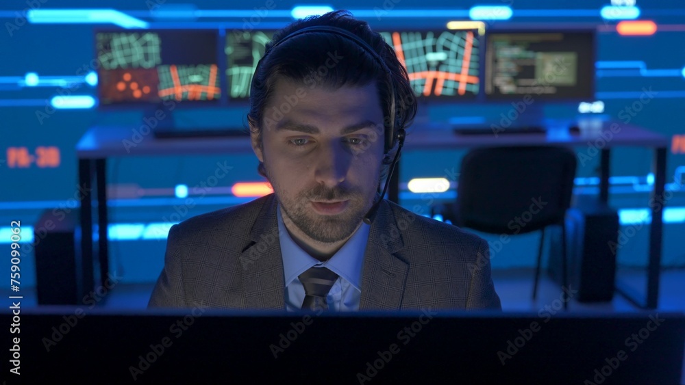 Technical support in control room. Man IT specialist works on computer talking in headset at the office, big digital screen shows infrastructure data. Technical support in control room. Man IT specialist works on computer talking in headset at the office, big digital screen shows infrastructure data.