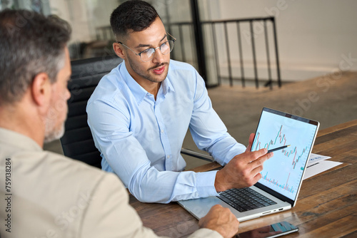 Financial advisor explaining invest stock market data consulting investor. Two busy business men analysts doing finance trading analysis pointing at exchange chart on laptop screen working in office.