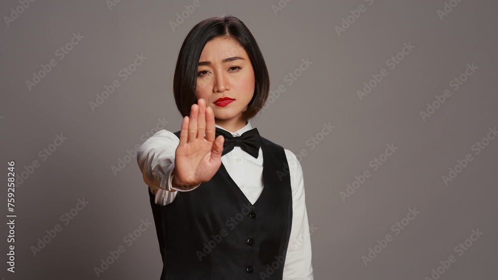 Asian administrator showing stop sign with palm raised in studio, presenting negative symbol and expressing disapproval. Woman receptionist doing no gesture, being displeased. Camera B. Asian administrator showing stop sign with palm raised in studio, presenting negative symbol and expressing disapproval. Woman receptionist doing no gesture, being displeased. Camera B.