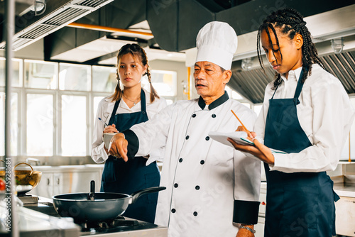 Asian senior chef in uniform educates multiracial students in a restaurant kitchen. Focus on teamwork learning and note-taking in this professional teaching environment. Food Edocation