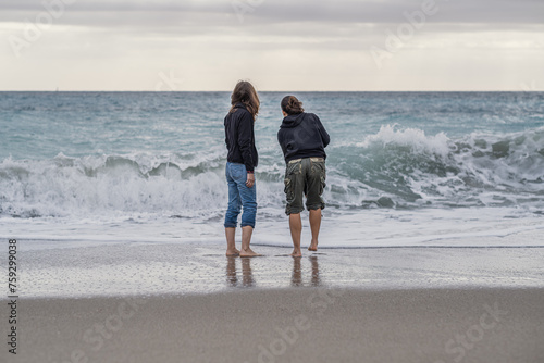 A mother and daughter standing on the beach, looking at the sea, rear view.