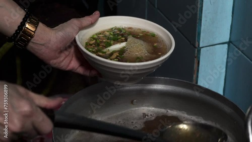 A close-up of hands of a person pouring meat and offal soup from a metal pot into a white ceramic bowl using a ladle. photo