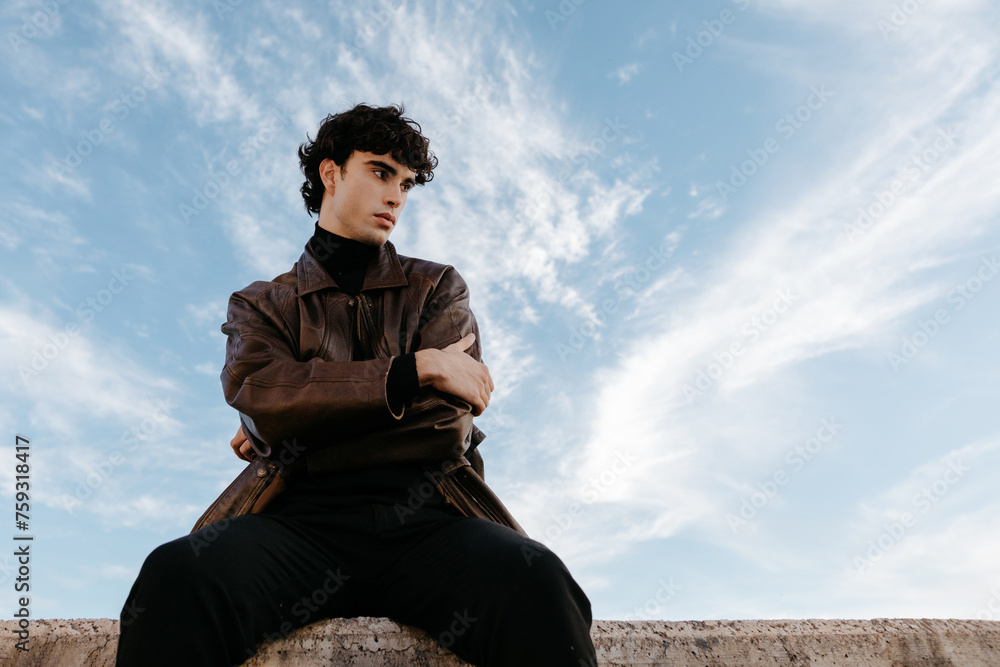 Stylish young man sitting on stone border against blue sky Stylish young man sitting on stone border against blue sky
