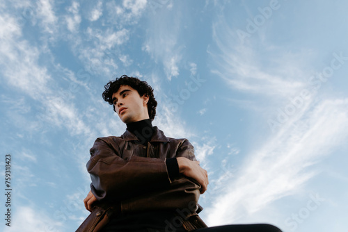 Young confident man in leather jacket against blue sky with clouds photo