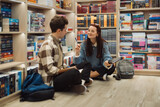 Two students engaged in discussion sitting on the library floor surrounded by bookshelves.
