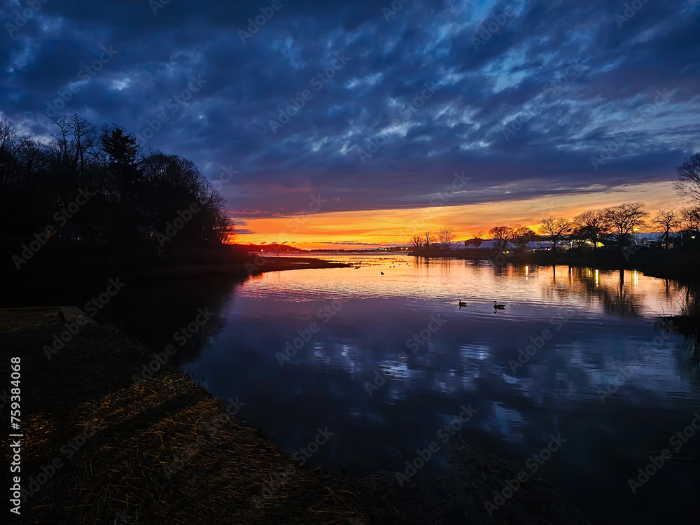 Captivating Long Island sunset landscapes over tranquil waters near Port Jefferson, showcasing a spectrum from blue to fiery orange hues under dramatic cloud-filled skies, reflecting in the serene riv