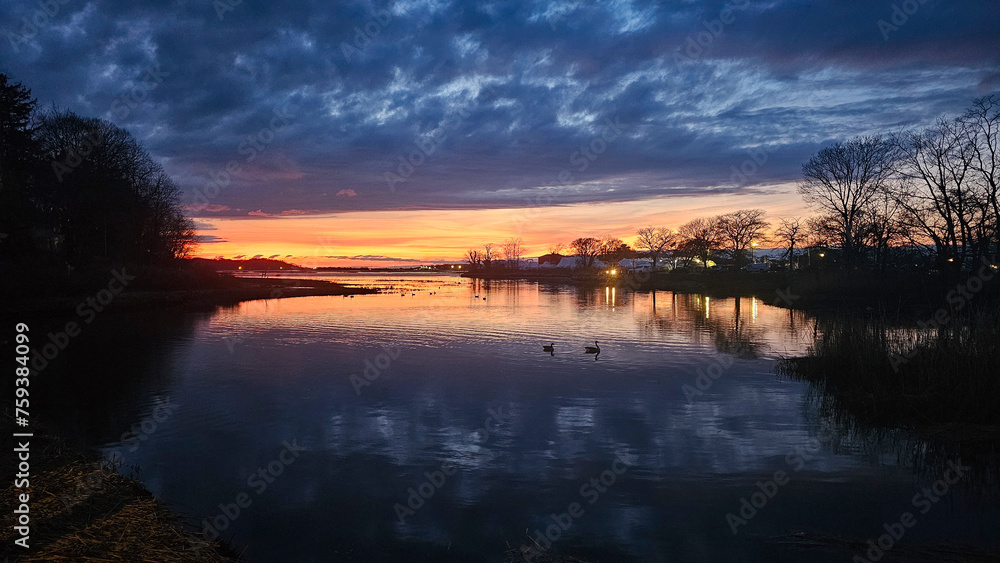Fototapeta premium Captivating Long Island sunset landscapes over tranquil waters near Port Jefferson, showcasing a spectrum from blue to fiery orange hues under dramatic cloud-filled skies, reflecting in the serene riv