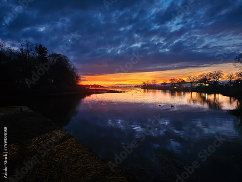 Captivating Long Island sunset landscapes over tranquil waters near Port Jefferson, showcasing a spectrum from blue to fiery orange hues under dramatic cloud-filled skies, reflecting in the serene riv