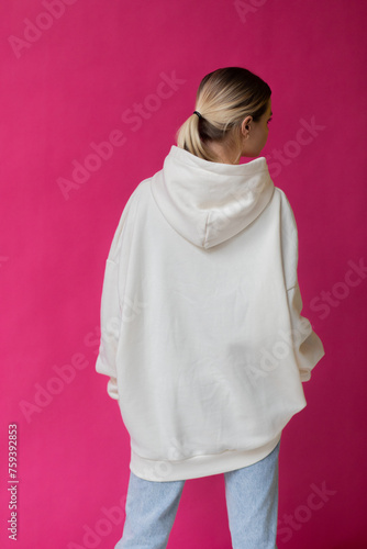 Beautiful blonde woman in a white hoodie and blue jeans posing on a pink background