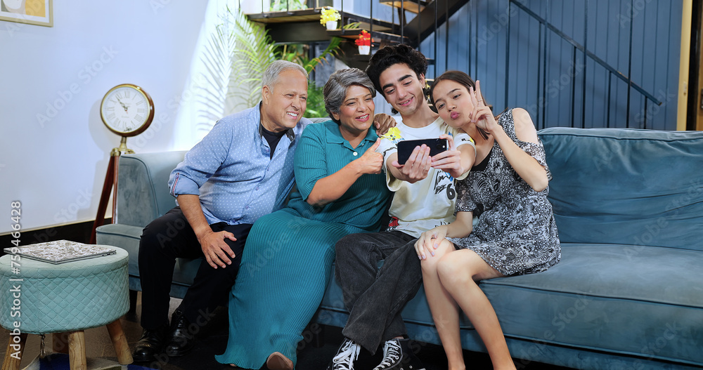 Fototapeta premium Indian Asian happy family group sitting sofa couch smiling boy man teen girl hold mobile use phone look cam take selfie home parents couple pose funny face photo record video vlog blog enjoy fun joy