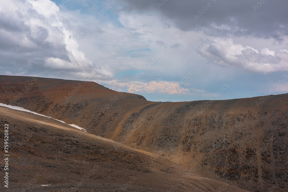 Sunlit large sharp ridge under gray rainy and sunset clouds in far away in changeable weather. Beautiful layered landscape with large mountain wall in sunlight and dramatic cloudy sky in sunset color.