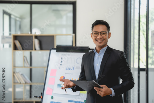 Asian businessman in a suit is standing in front of a white board with a presentation. © amnaj