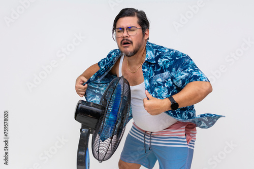 Obraz na plátně A desperate middle-aged man in an unbuttoned Hawaiian shirt feeling the heat, cooling off in front of an electric fan, isolated on white background