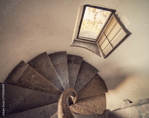Old stone staircase in a tower - view from above at the stairwell. Perspective looking down view of clockwise round-shaped staircase. Small window overseeing the garden.