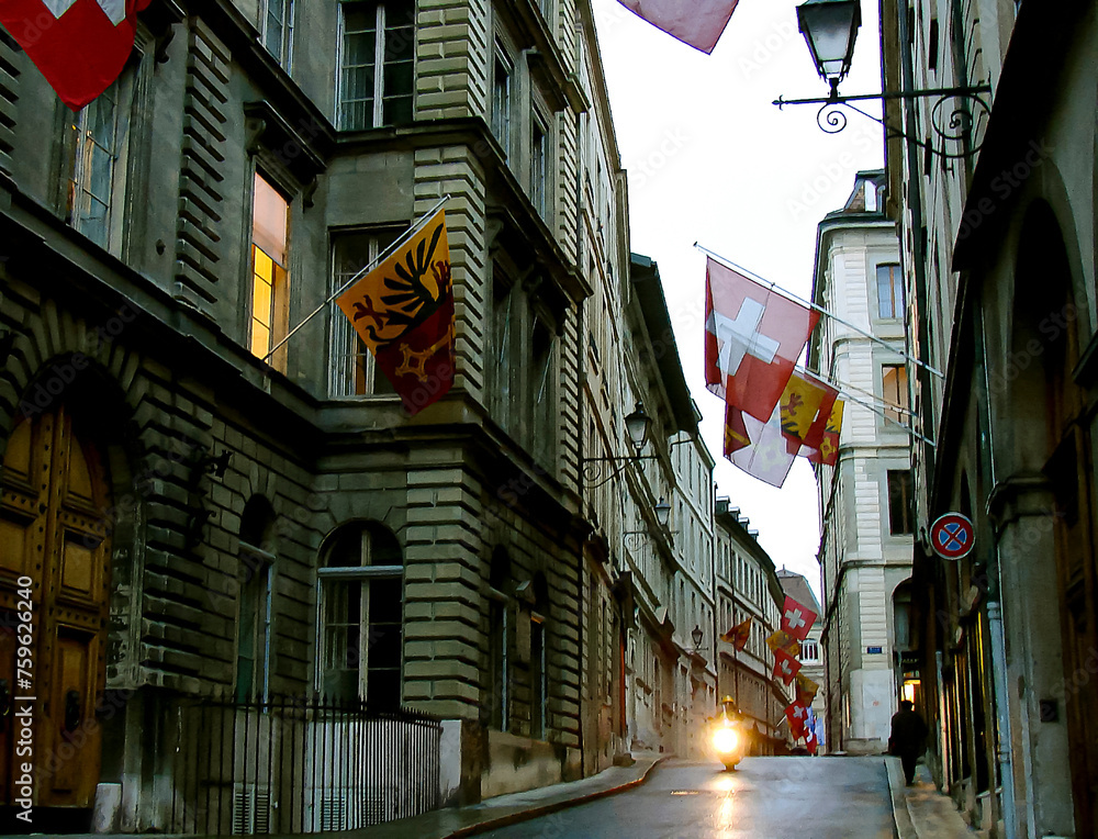 Fototapeta premium evening Geneva: quiet narrow European street; mid-rise buildings; Swiss flags; a lonely old man walks off into the distance; one policeman rides a motorcycle with his headlights on