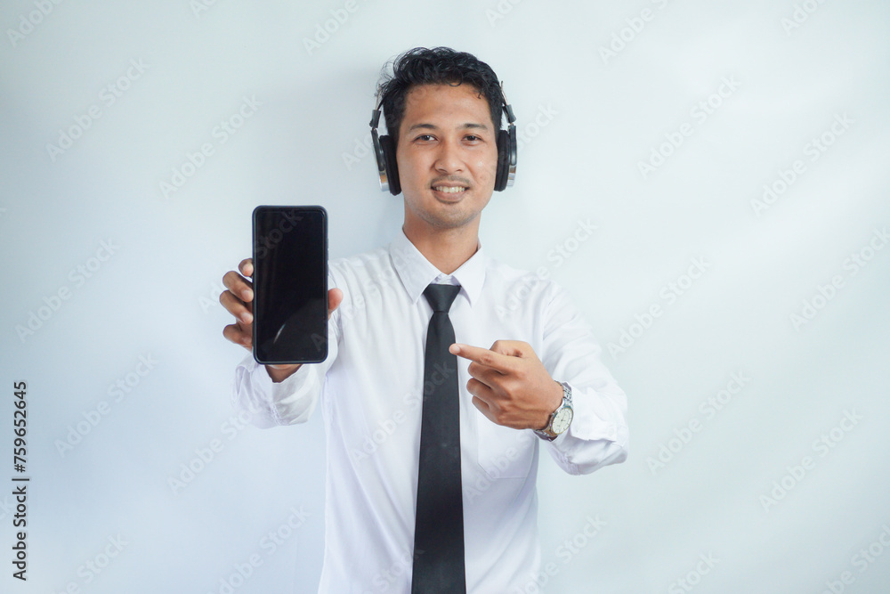 Adult Asian man smiling confident while pointing finger to blank handphone screen. using headphone Adult Asian man smiling confident while pointing finger to blank handphone screen. using headphone
