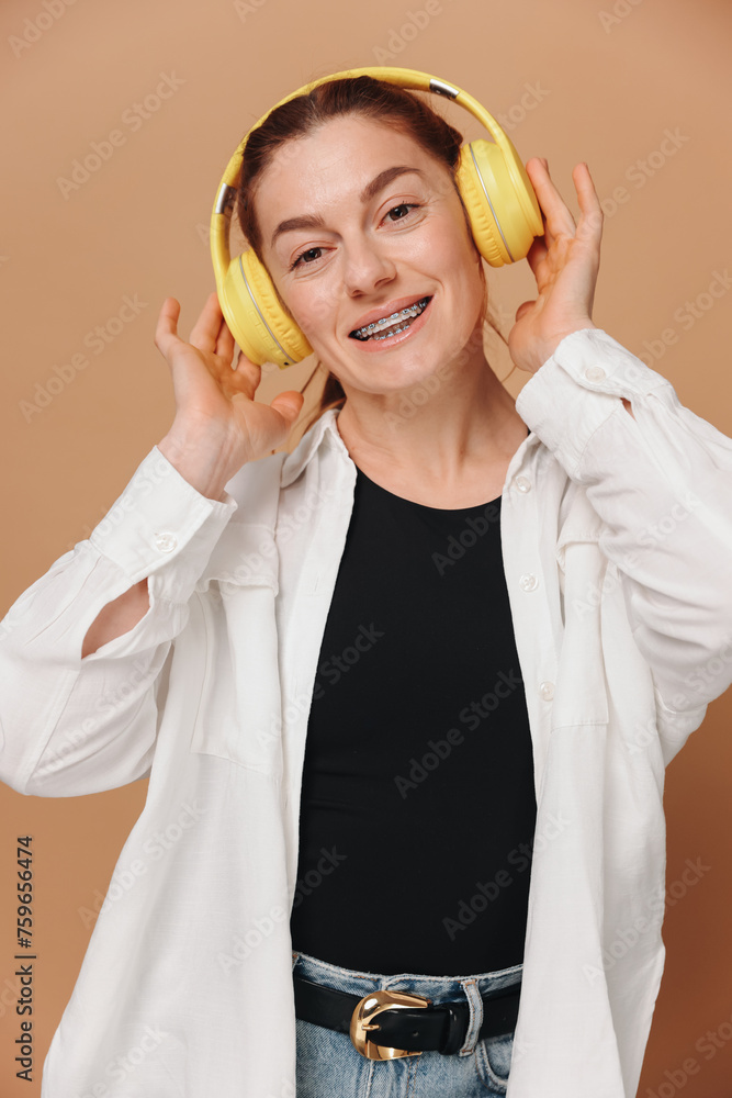 Modern woman smiling with braces on her teeth and listening to music in headphones on a beige background Modern woman smiling with braces on her teeth and listening to music in headphones on a beige background