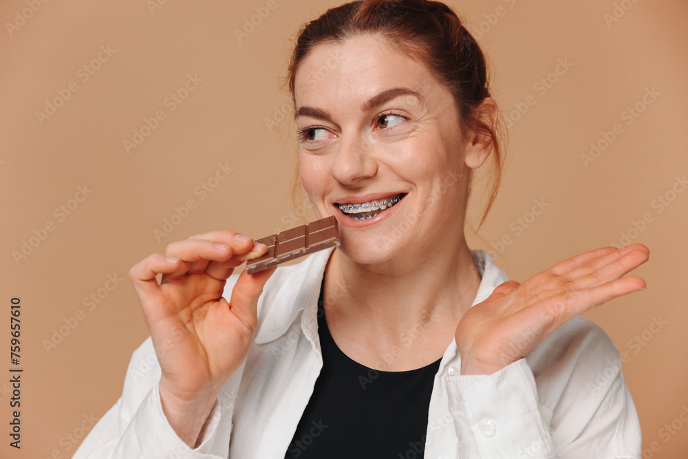 Portrait of mature woman in braces biting chocolate on a beige background