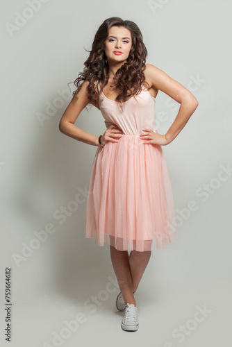 Glorious fashion model woman in pink dress and white shoes on feet standing against white studio wall background