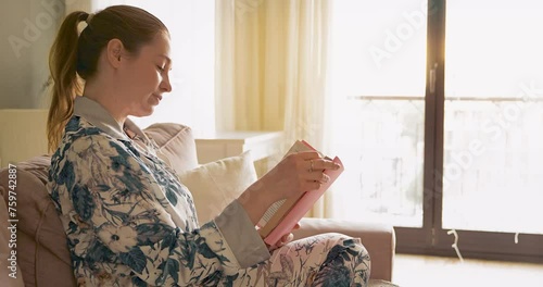 a woman is reading a book at home on a sofa in the beautiful daylight.