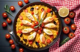 Top view of paella with seafood, tomatoes and herbs in a frying pan on the table with white and red checkered napkin and cutlery