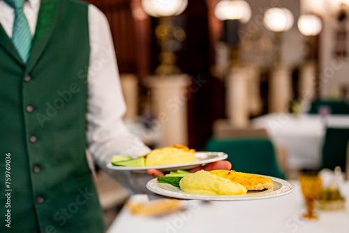 Waiter in green vest serving two plates of delicious meals in an upscale restaurant with elegant decor and lighting