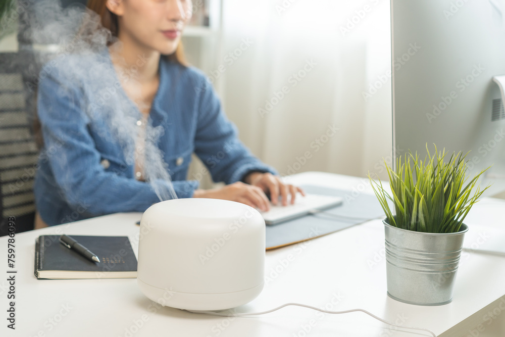 Modern air humidifier device during sitting workplace, happy asian young woman using computer work on internet, enjoying aromatherapy steam scent from essential oil diffuser in room at home office. - obrazy, fototapety, plakaty 
