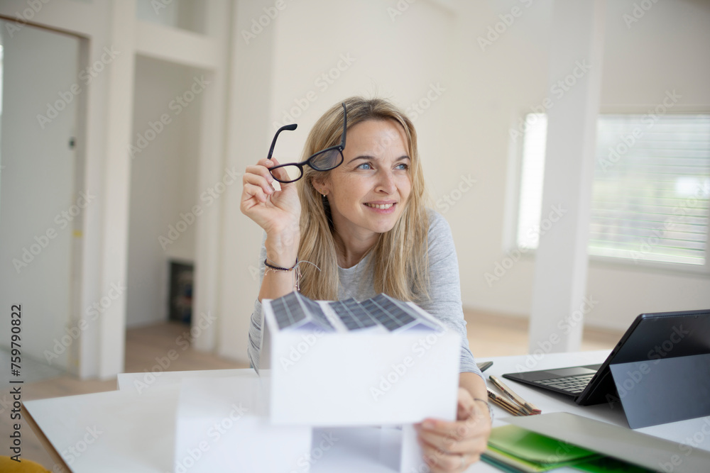 pretty blonde architect with black eyeglasses is standing at a desk and looking at a model of a house with photovoltaic system, solar system, in a loft, office pretty blonde architect with black eyeglasses is standing at a desk and looking at a model of a house with photovoltaic system, solar system, in a loft, office