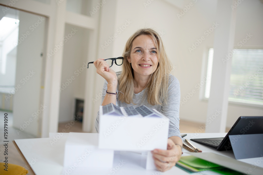 pretty blonde architect with black eyeglasses is standing at a desk and looking at a model of a house with photovoltaic system, solar system, in a loft, office pretty blonde architect with black eyeglasses is standing at a desk and looking at a model of a house with photovoltaic system, solar system, in a loft, office