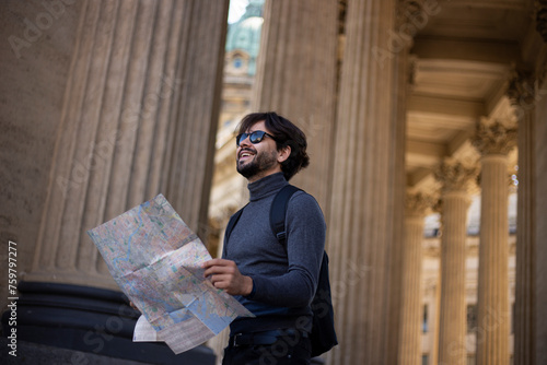 Happy young man looking up, holding a map, trying to find a destination, isolated historical city with columns.
