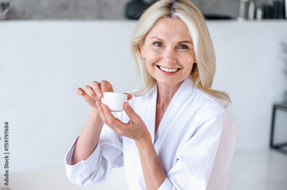 50 years old woman in white bathrobe holding jar with moisturizer cream 50 years old woman in white bathrobe holding jar with moisturizer cream