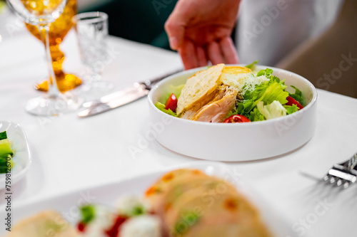 A fresh salad with grilled chicken, greens, and cherry tomatoes served at a well-set dining table. Utensils neatly placed beside the plates, and two glasses—one with an orange beverage—are visible.