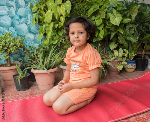 Little Girl Child smiling sitting and relaxing after practicing Yoga on red mat at Terrace Garden of House. Wellness and healthy lifestyle of children
