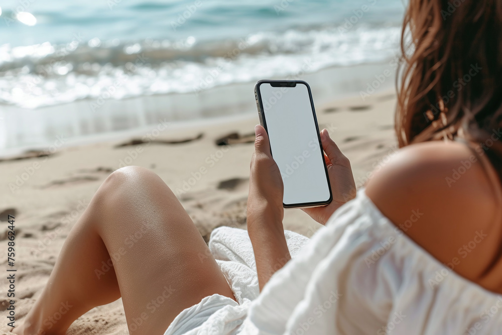 Mockup image of a woman holding and using mobile phone with blank desktop screen while sitting on the beach