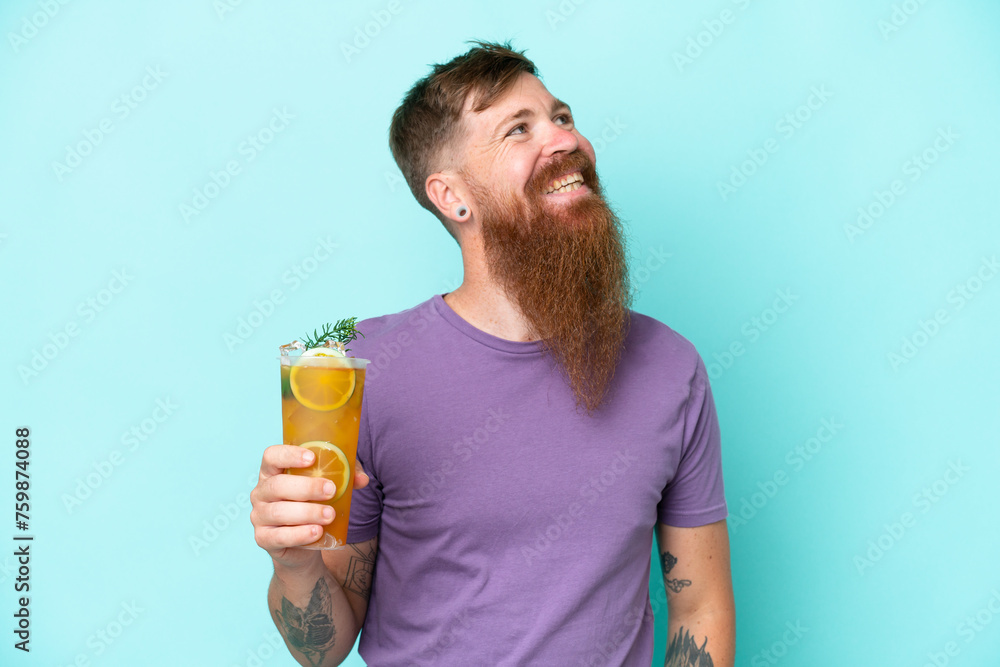Redhead man with long beard holding a cocktail isolated on blue background looking up while smiling Redhead man with long beard holding a cocktail isolated on blue background looking up while smiling