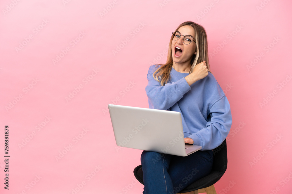 Young woman sitting on a chair with laptop over isolated pink background celebrating a victory Young woman sitting on a chair with laptop over isolated pink background celebrating a victory