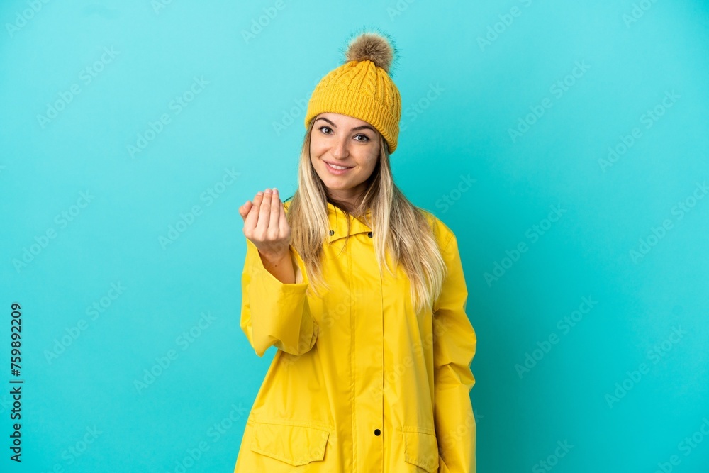Young woman wearing a rainproof coat over isolated blue background inviting to come with hand. Happy that you came Young woman wearing a rainproof coat over isolated blue background inviting to come with hand. Happy that you came