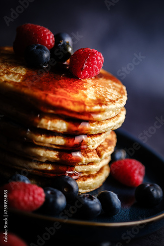 Stacked pancakes with raspberries and blueberries and maple syrup against a dark background.
