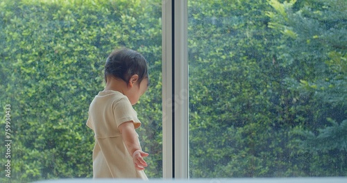 A young child is standing in front of a window, looking out at the trees. Scene is peaceful and contemplative, as the child seems to be lost in thought
