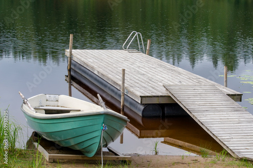 Wooden dock and a green rowing boat anchored at shore. Lake at background with trees reflecting from the water.