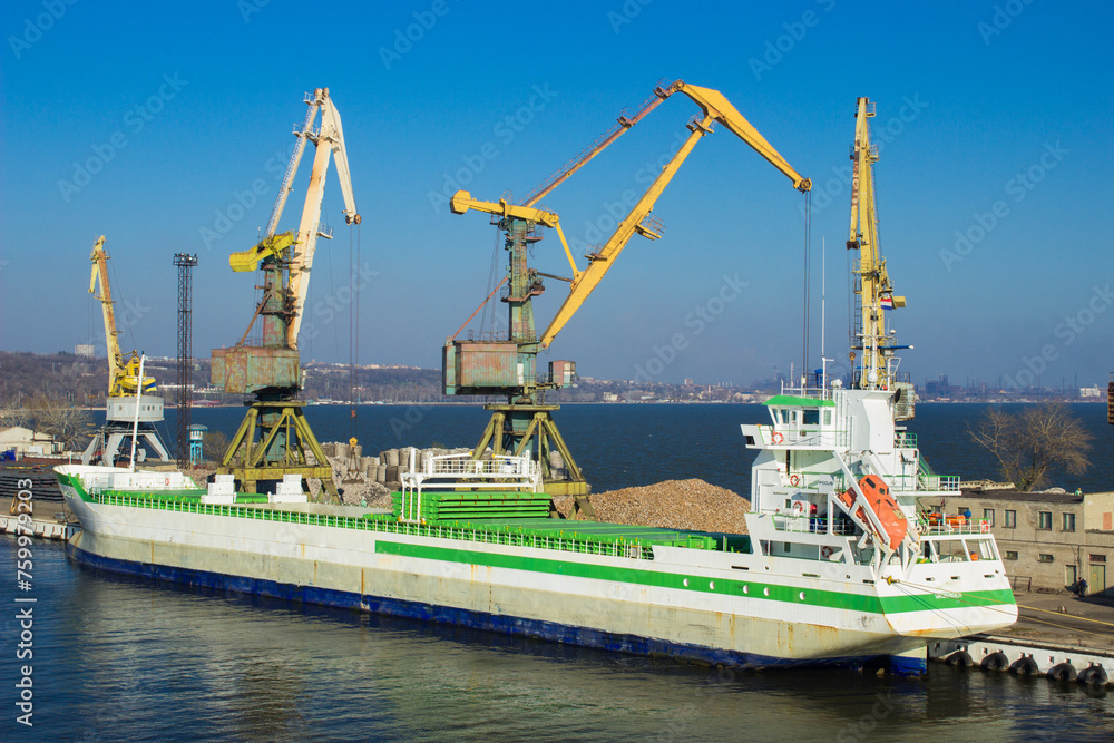 Cargo ship docked at international port, cranes loading containers under clear sky. Maritime freight transport, global trade, logistics network. Import, export operations in coastal facility.